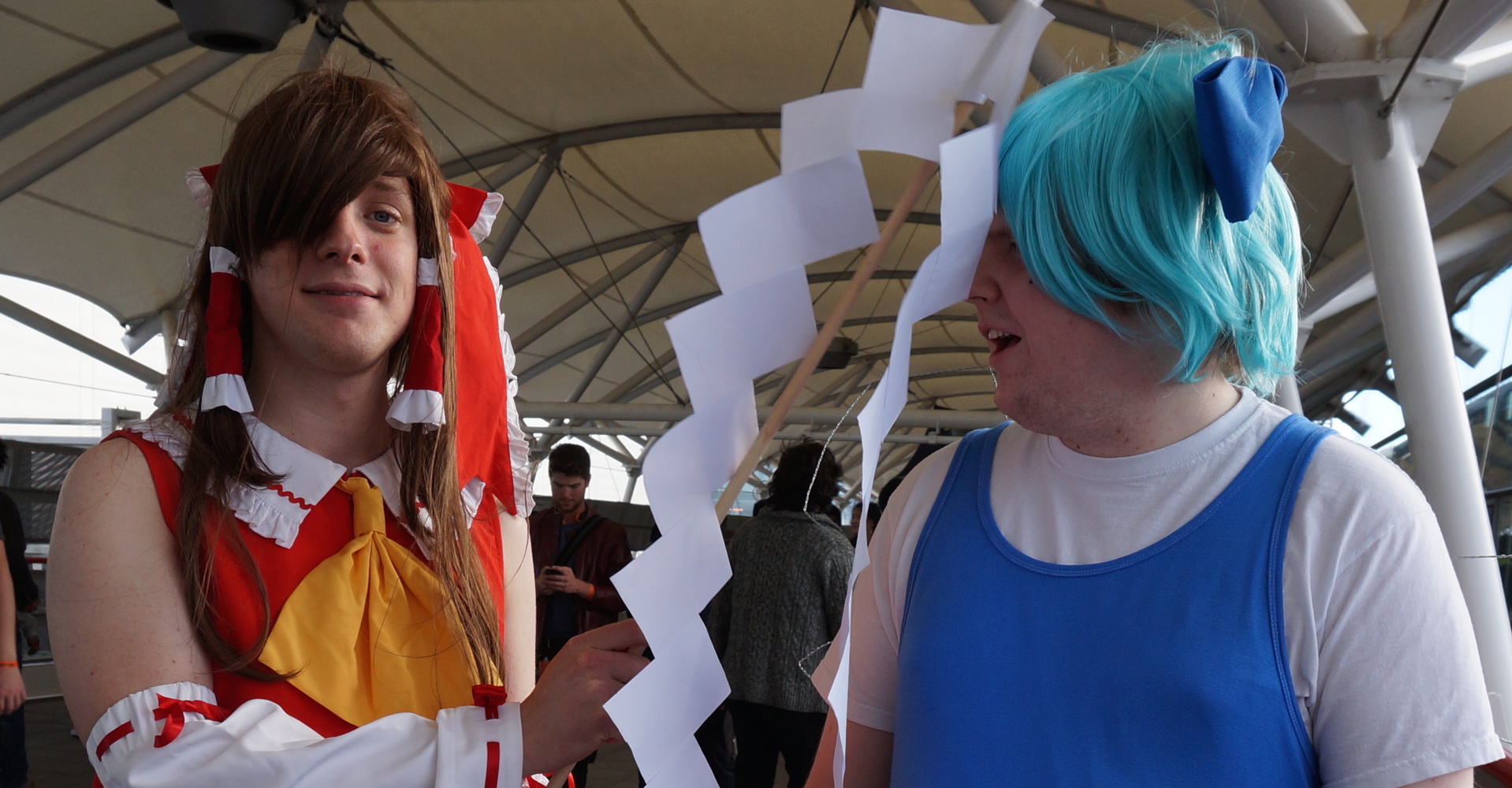 Two white dudes dressed as Touhou characters. The one on the left (Reimu, in a frilly red dress and long brown wig) is hitting the one on the right (Cirno, wearing a short blue wig, a blue dress, and wire fairy wings) with a home-made gohei.